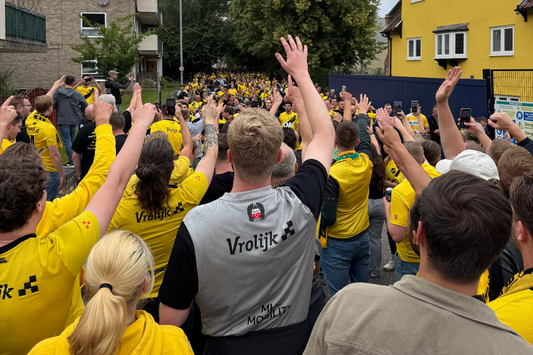 Een grote groep NAC Breda supporters in gele shirts en hoodies loopt door een straat, zwaaiend met hun armen in de lucht, op weg naar het stadion in Norwich. Op de achtergrond is een geel gebouw te zien.