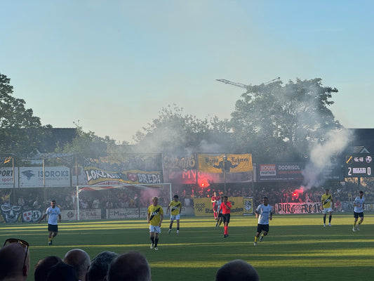 feerimpressie van de oefenwedstrijd tussen NAC Breda en Lokeren op 10 juli 2025, met spelers op het veld en fans op de tribune die spandoeken tonen en fakkels afsteken, wat zorgt voor een dichte rookontwikkeling.