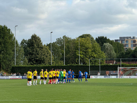 De spelers van het Zeeuws elftal in blauwe tenues en NAC Breda in gele en zwarte tenues staan in een kring op het voetbalveld voorafgaand aan de oefenwedstrijd op 15 juli 2025. Op de achtergrond zijn bomen, gebouwen en reclameborden zichtbaar