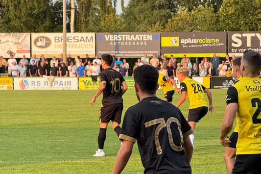 Spelers van NAC Breda in zwarte shirts met gele accenten tijdens een oefenwedstrijd tegen OFI Kreta op 24 juli 2024, gezien vanaf achter het doel met supporters op de tribune en reclameborden op de achtergrond.