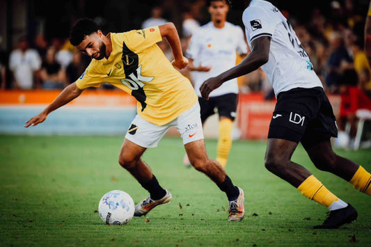 Foto van Vinnie de Laat. Speler van NAC Breda in een geel-zwart tenue aan de bal, dribbelend langs een speler van Lokeren in een wit tenue, tijdens de oefenwedstrijd op 10 juli 2025.