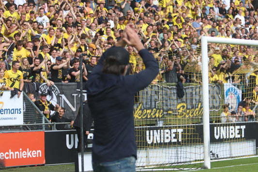 NAC trainer Carl Hoefkens staat op het voetbalveld, van achteren gefotografeerd, terwijl hij kijkt naar een tribune vol supporters en rook.