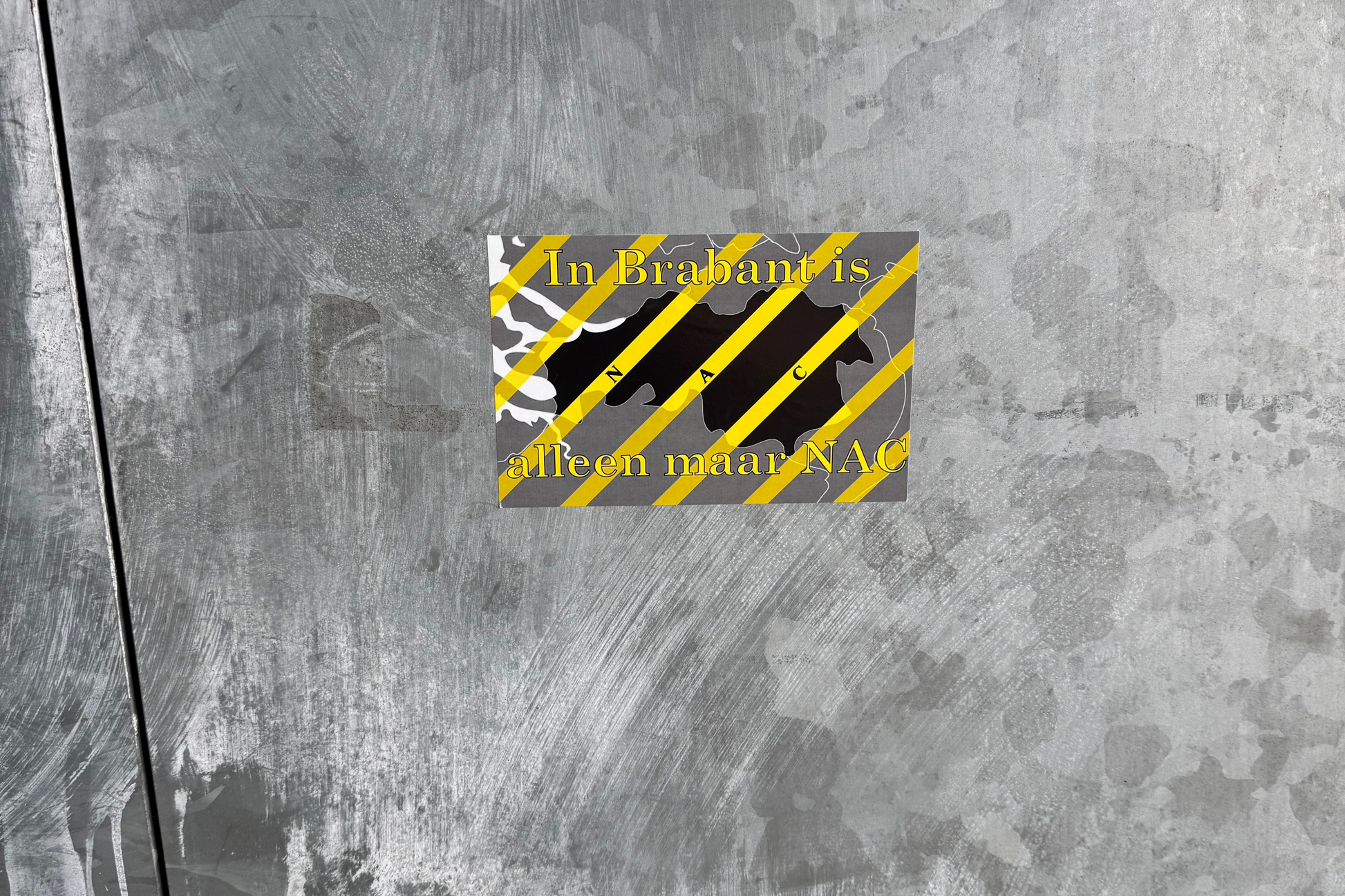 Een 'In Brabant is alleen maar NAC' sticker geplakt op een ruwe, grijze metalen ondergrond, bij het stadion. De sticker toont de omtrek van Noord-Brabant in zwart met gele diagonale strepen en de letters 'N A C', omringd door dezelfde gele strepen op een grijze achtergrond. Rondom de sticker zijn gebruikssporen en vegen te zien op het metaal.