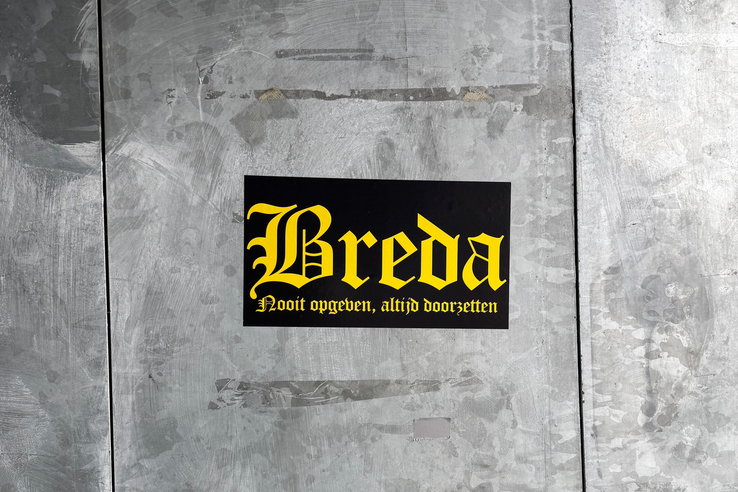 Een zwarte 'Breda' sticker met gele gotische letters en de tekst 'Nooit opgeven, altijd doorzetten' eronder, geplakt op een lichtgrijze, robuuste metalen ondergrond met zichtbare vegen en krassen, bij het NAC Breda stadion.