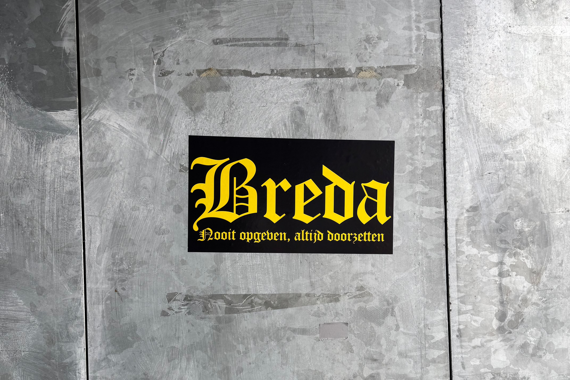 Een zwarte 'Breda' sticker met gele gotische letters en de tekst 'Nooit opgeven, altijd doorzetten' eronder, geplakt op een lichtgrijze, robuuste metalen ondergrond met zichtbare vegen en krassen, bij het NAC Breda stadion.