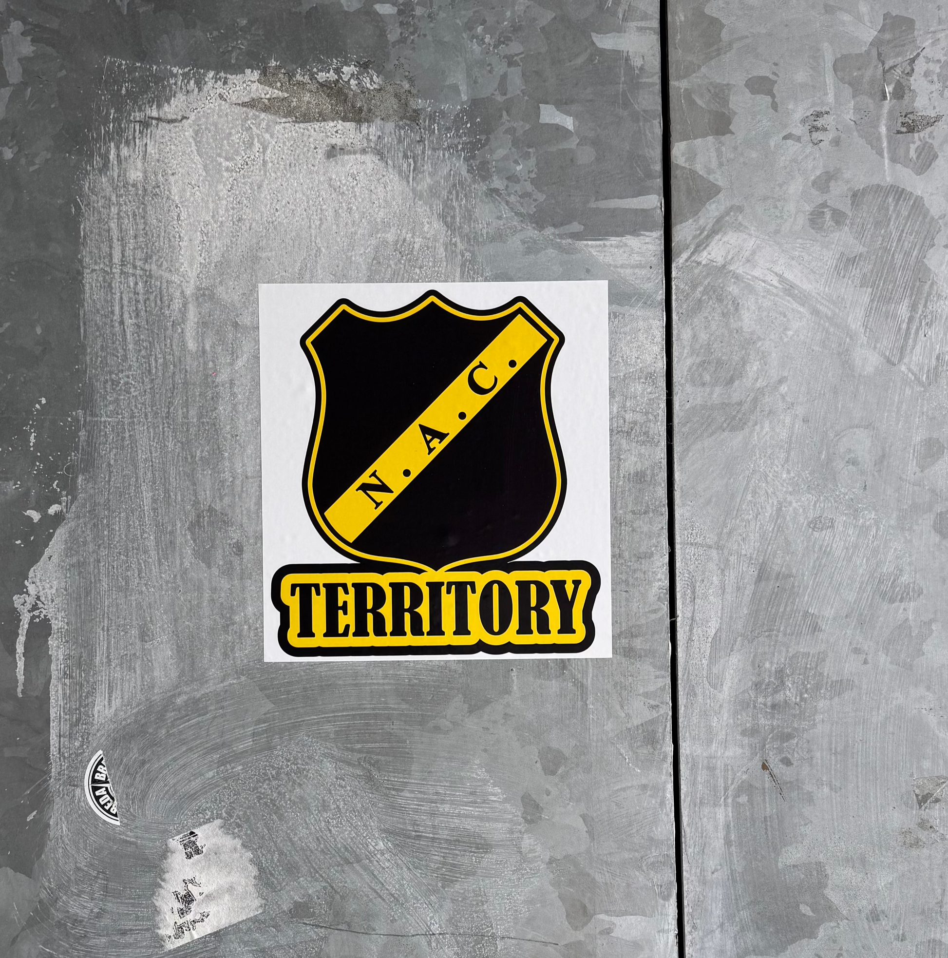 Een 'NAC Territory' sticker geplakt op een ruwe, grijze metalen plaat, typerend voor de omgeving van een stadion. De sticker toont het NAC Breda logo, een zwart schild met gele rand en een diagonale gele balk met de letters 'N.A.C.', en daaronder in gele letters met zwarte omranding 'TERRITORY'. De metalen ondergrond vertoont krassen en gebruikssporen.