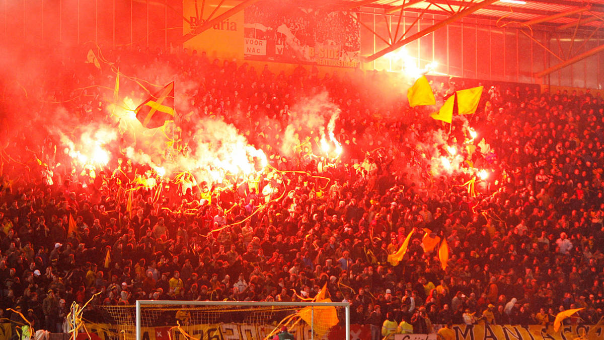 Deze foto legt de intense en gepassioneerde sfeer vast in het Rat Verlegh Stadion tijdens een voetbalwedstrijd van NAC Breda. De foto toont een grote menigte fans die rode fakkels hebben aangestoken, waardoor het stadion gehuld is in een dikke, rode rook. De gele en zwarte kleuren van NAC Breda zijn prominent aanwezig in de vlaggen en kleding van de fans. De foto toont de passie en toewijding van de NAC Breda-fans en de unieke sfeer in het stadion.