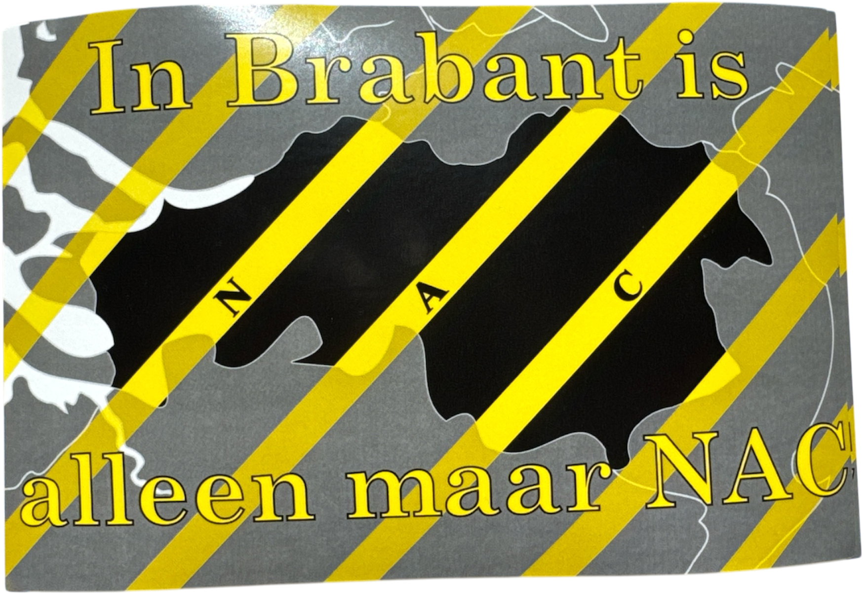 "Rechthoekige sticker met een grijze achtergrond en gele diagonale strepen, lijkend op waarschuwingstape. Centraal staat een zwarte omtrek van de provincie Noord-Brabant met daaroverheen gele diagonale strepen en de letters 'N A C'. Bovenaan staat in gele letters 'In Brabant is' en onderaan 'alleen maar NAC', afmetingen 16x8cm.