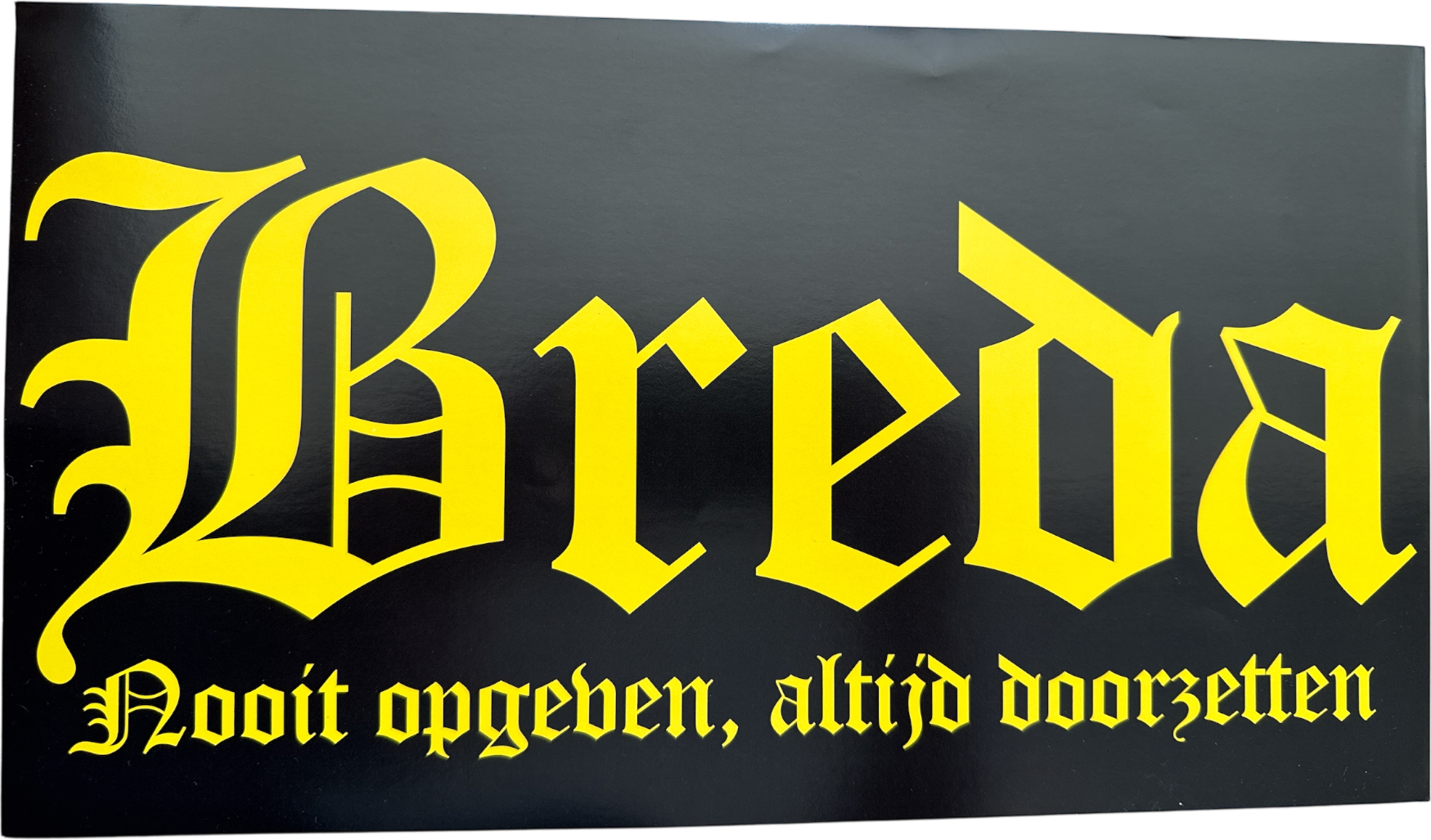 Zwarte sticker met in geel gotisch schrift 'Breda' en daaronder de tekst 'Nooit opgeven, altijd doorzetten' in hetzelfde gele lettertype, afmetingen 36x20cm.