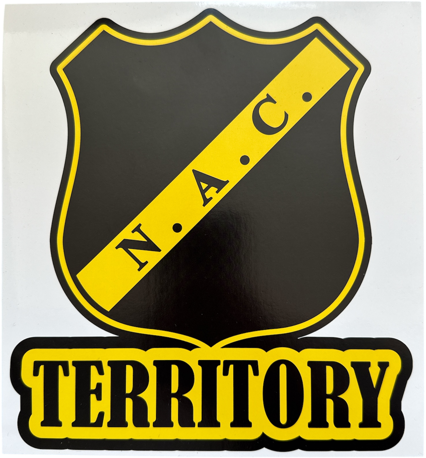Zwart schildvormige NAC Breda sticker met gele rand en een gele diagonale band met daarin de zwarte letters 'N.A.C.'. Daaronder staat in gele letters met zwarte omranding het woord 'TERRITORY', afmetingen 22x22cm.