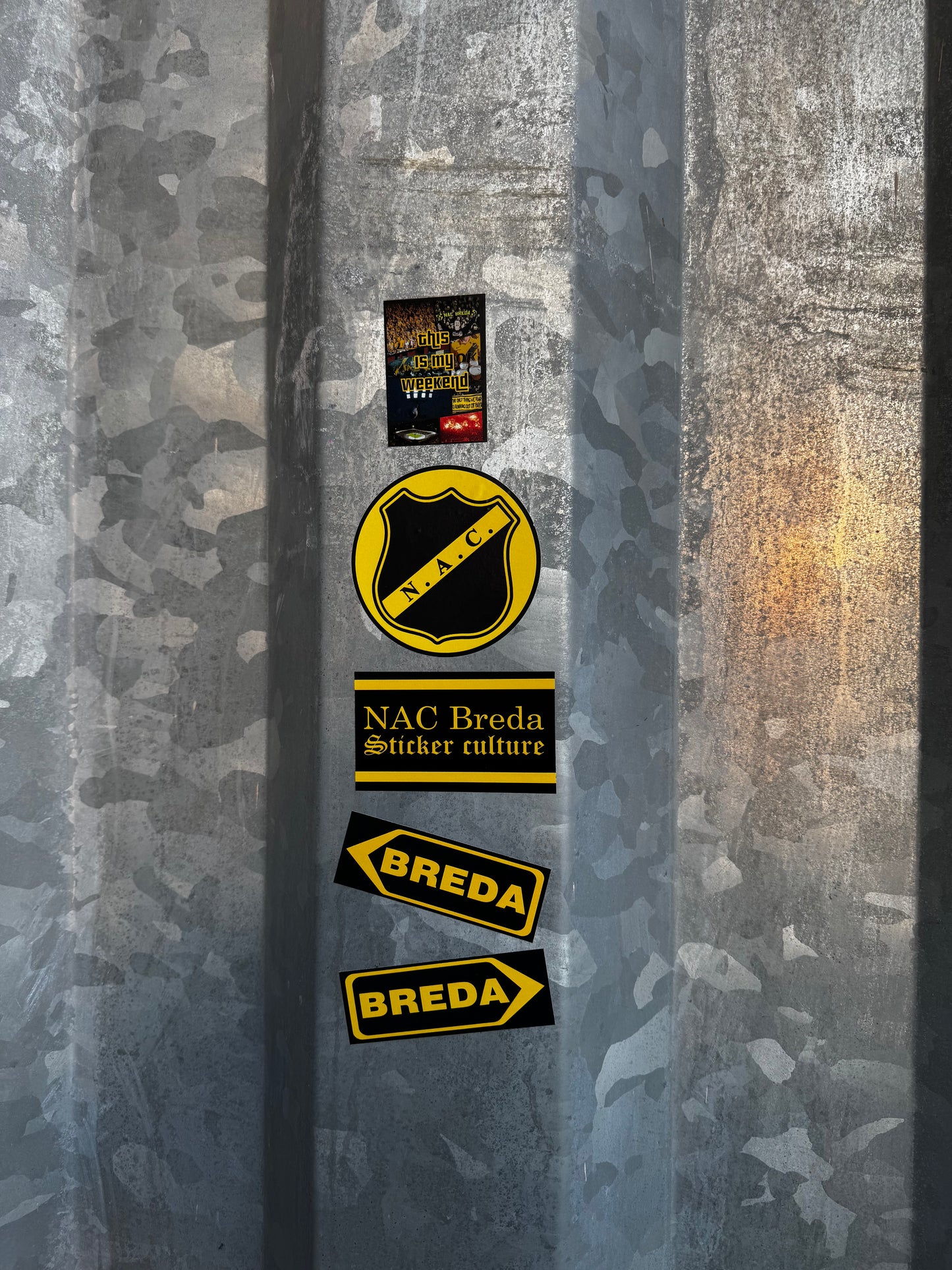 NAC Breda stickers uit pakket 1, waaronder het officiële logo en Breda richtingaanwijzers, aangebracht op de ruwe, gegalvaniseerde hekken van het NAC stadion.