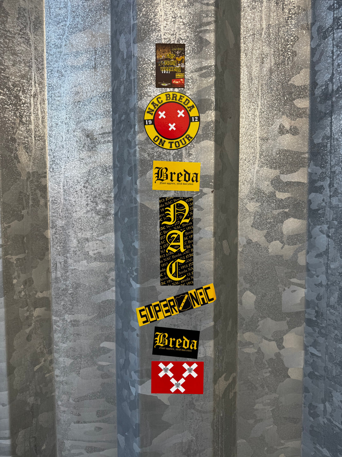 Overzicht van diverse NAC Breda stickers, waaronder NAC On Tour, Super NAC en de leus Nooit Opgeven Altijd Doorzetten, geplakt op de karakteristieke gegalvaniseerde stadionhekken rond het Rat Verlegh Stadion.