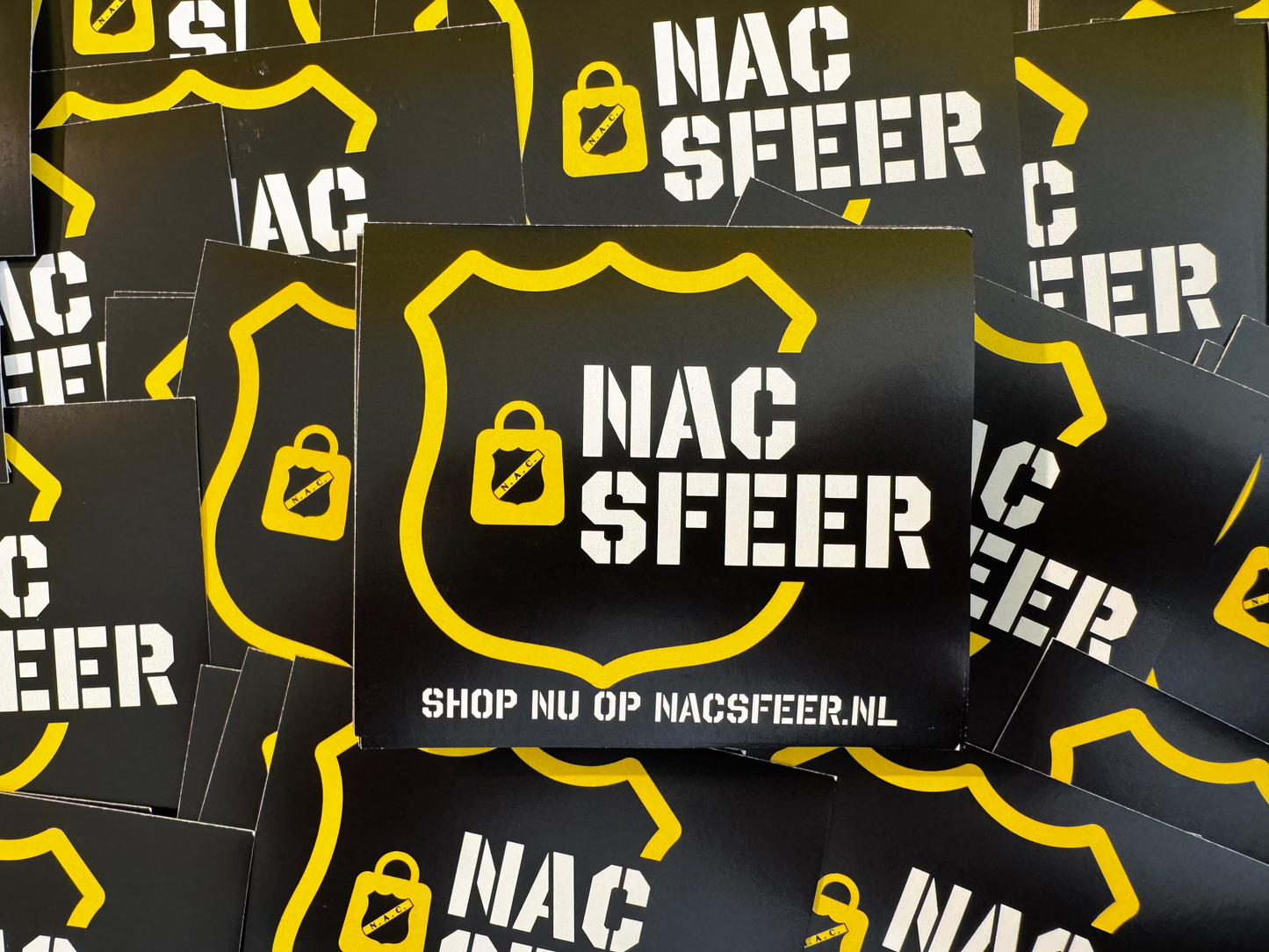 Stapel zwarte promotiestickers met het gele NACsfeer logo, het slotje icoon en de webshoptekst 'Shop nu op NACsfeer.nl'.