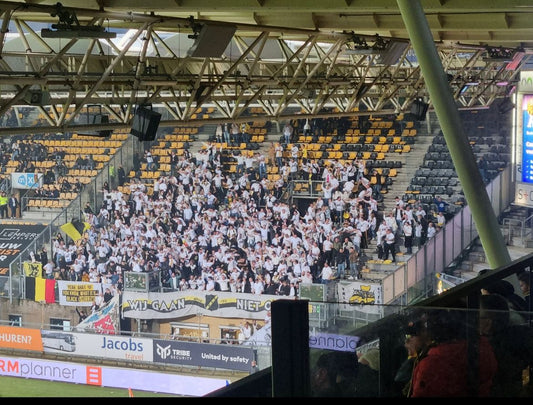 Een breed overzicht van een voetbalstadion gevuld met een grote groep NAC Breda supporters gekleed in het wit, juichend op de tribune. Foto van het uitvak bij Roda in Kerkrade, NAC won de wedstrijd met 0-5 in de play-offs voor promotie