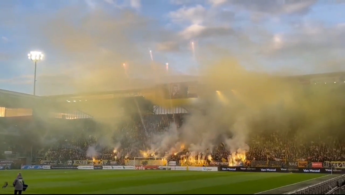 Een voetbalstadion gevuld met dichte gele en witte rook, afkomstig van fakkels die branden op de tribunes. De rook belemmert het zicht op de toeschouwers, maar de contouren van de tribunes en de verlichting van het stadion zijn zichtbaar.