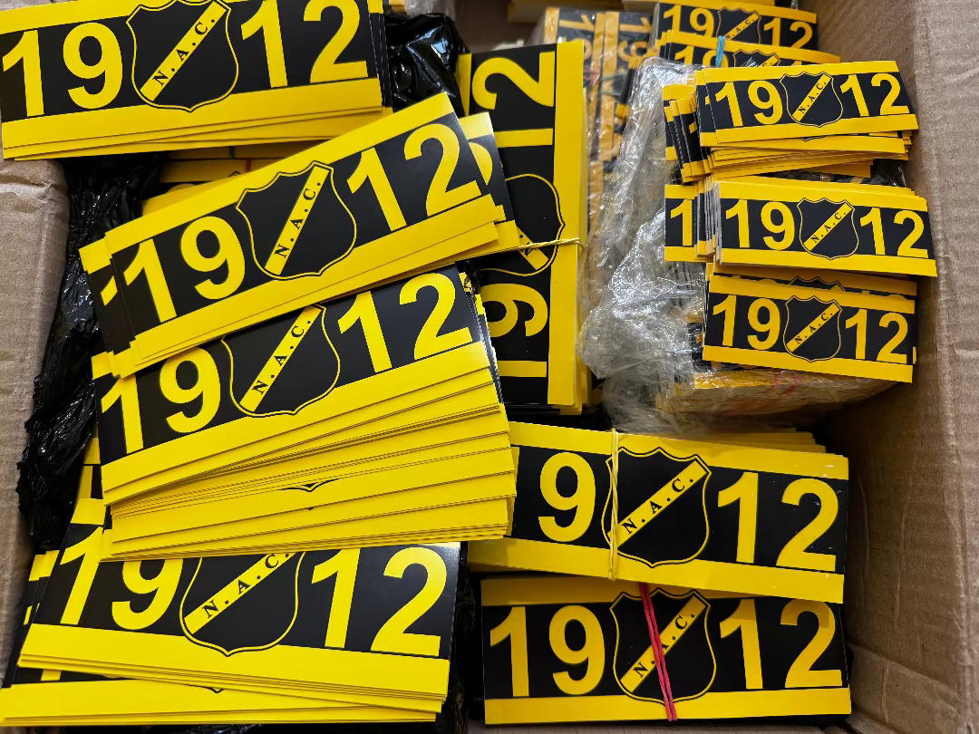 Geel-zwarte NAC Breda 1912 stickers opgestapeld in een doos, voorzien van het officiële NAC logo.