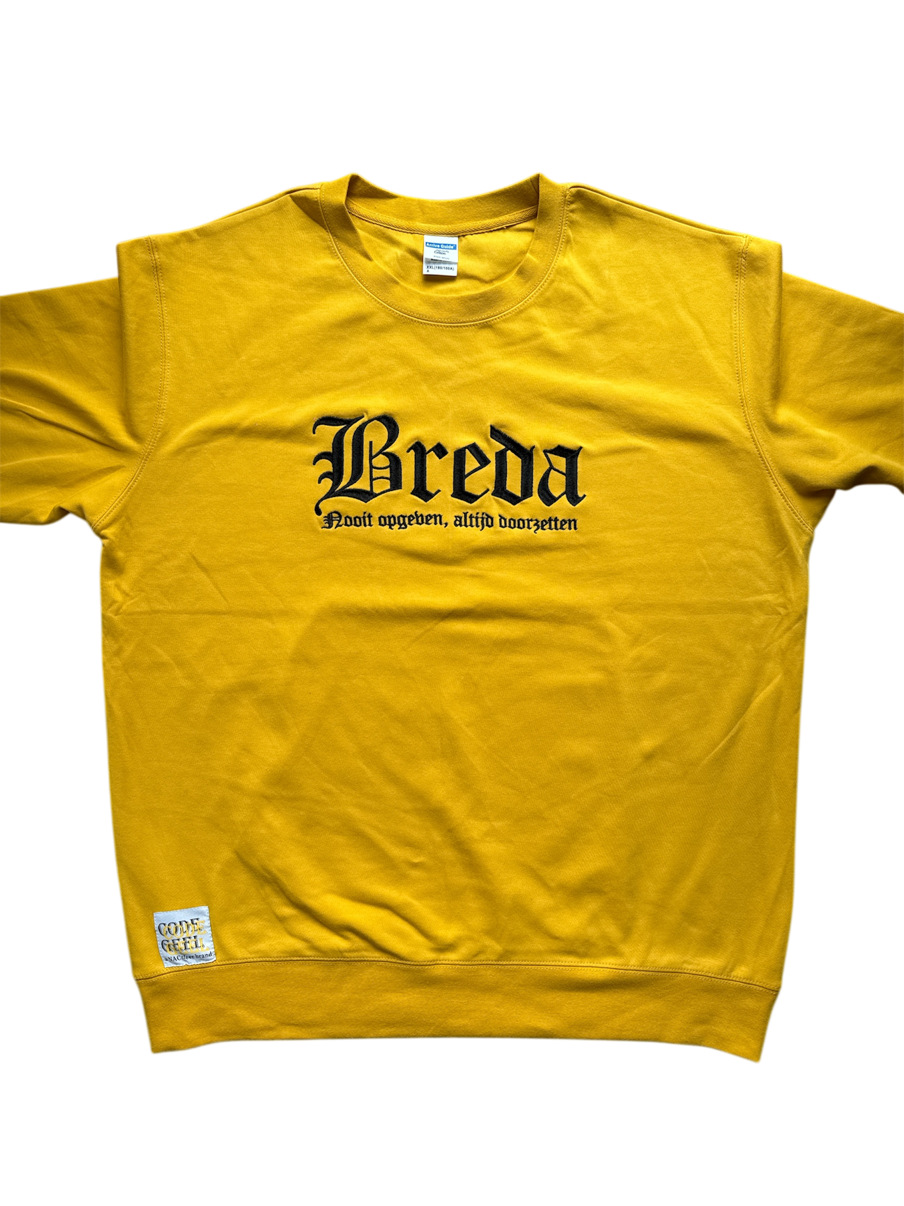 Gele trui met in zwarte, gotische letters de tekst 'Breda' en daaronder in een kleiner lettertype 'Nooit opgeven, altijd doorzetten'. Linksonder is een wit label met 'CODE GEEL' bevestigd. De trui ligt plat op een lichtgrijze achtergrond.