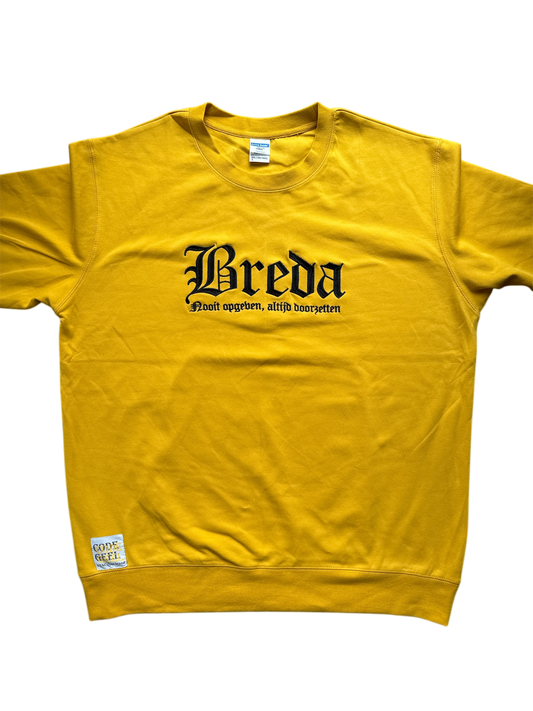 Gele trui met in zwarte, gotische letters de tekst 'Breda' en daaronder in een kleiner lettertype 'Nooit opgeven, altijd doorzetten'. Linksonder is een wit label met 'CODE GEEL' bevestigd. De trui ligt plat op een lichtgrijze achtergrond.