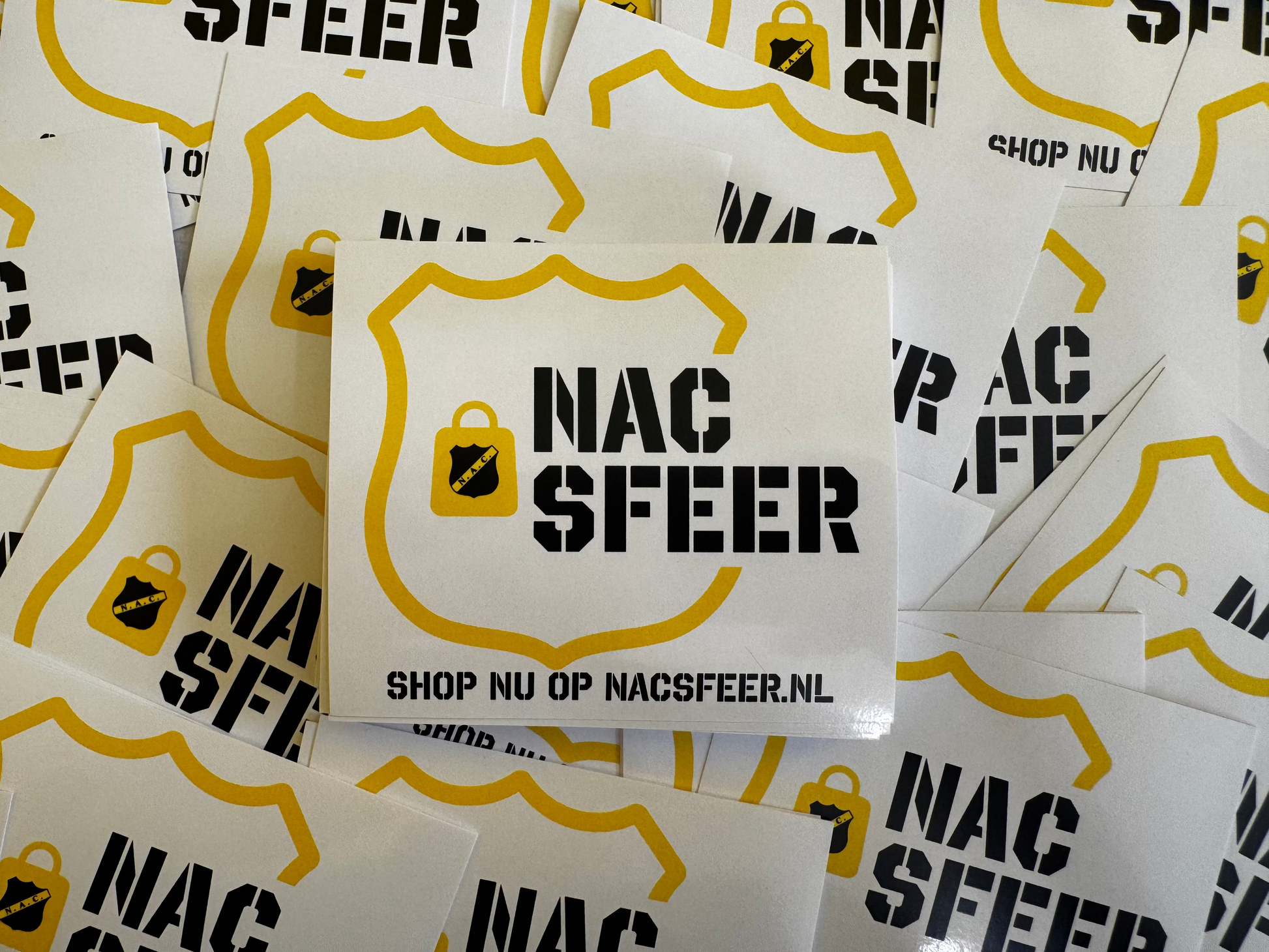 Stapel witte promotiestickers met het zwarte en gele NACsfeer logo, het slotje icoon en de webshoptekst 'Shop nu op NACsfeer.nl'.